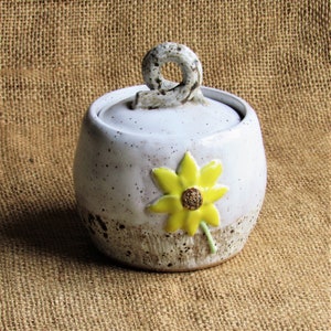 May include: A white ceramic jar with a brown speckled glaze and a yellow flower design. The jar has a lid with a unique handle.