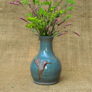 May include: A teal ceramic vase with a hummingbird design, holding a bouquet of green and purple flowers. The vase has a speckled finish and a flared rim. The hummingbird is brown with detailed wing and beak features.