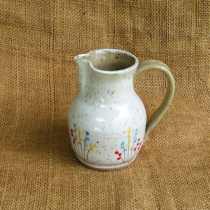 May include: A handmade ceramic pitcher with a cream-colored glaze and speckled finish. The pitcher features a curved handle and a spout for pouring. Delicate floral designs in red, yellow, and blue adorn the lower portion of the pitcher, adding a touch of color.
