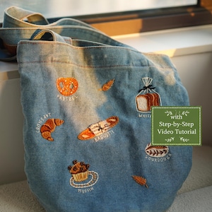Op de afbeelding: Een lichtblauwe denim tas met geborduurde afbeeldingen van een krakeling, croissant, stokbrood, witbrood, zuurdesem en muffin, elk voorzien van een label. Een groen rechthoekig label bevat de tekst with Step-by-Step Video Tutorial.