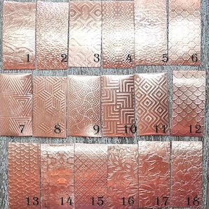 May include: Eighteen rectangular copper metal pieces, each with a unique embossed pattern. The pieces are numbered 1 to 18 and arranged on a wooden surface. The patterns include geometric shapes, textures, and organic designs.