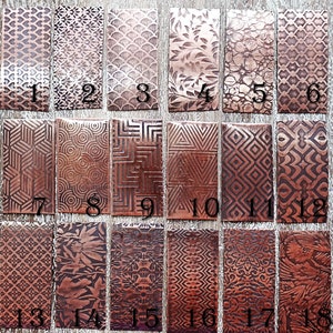 May include: Eighteen rectangular copper metal pieces with various embossed patterns, including floral, geometric, and abstract designs. Each piece is numbered from 1 to 18. The pieces are arranged on a light brown wooden surface.