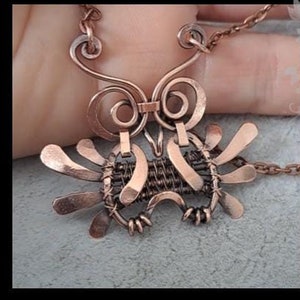 Może przedstawiać: Naszyjnik z zawieszką w kształcie sowy owiniętej miedzianym drutem. Sowa ma duże oczy i szczegółowe ciało. Naszyjnik jest na łańcuszku i jest trzymany przez rękę. Tekst "DIY Wire wrap baby Owl by Gemseeker Studio" znajduje się w tle.