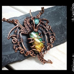 Może przedstawiać: Wisiorek w kształcie smoka owinięty miedzianym drutem z zielonym i żółtym kamieniem szlachetnym. Wisiorek leży na czarnej powierzchni łupka. DIY WIRE WRAPPED DRAGON BY GEMSEEKER STUDIO.