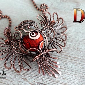 Może przedstawiać: Naszyjnik DIY z zawieszką w kształcie ptaka z drutu miedzianego z czerwonym kamieniem szlachetnym. Zawieszka wykonana jest z drutu owiniętego wokół czerwonego kamienia szlachetnego. Naszyjnik leży na drewnianej powierzchni.