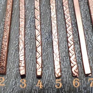 May include: Close-up of eight copper hair sticks with various embossed patterns. The hair accessories are numbered 1 to 8. The designs include floral, geometric, and linear motifs. The hair sticks are displayed on a textured, grey surface.