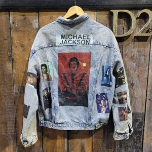 May include: A light blue denim jacket with various patches featuring images of Michael Jackson. The patches include a large image of Michael Jackson in a red and black outfit, a smaller image of Michael Jackson in a white shirt and black tie, and a smaller image of Michael Jackson in a black and white outfit. The jacket also has a patch with the text "MICHAEL JACKSON" on it.