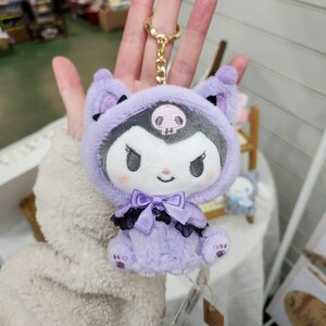 May include: Purple plush keychain with a black and white character wearing a purple hooded cape with a skull design. The character has a black bow and a white face with black eyes and a pink nose.