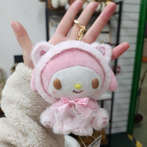 May include: Pink plush keychain featuring a character with a white face, brown eyes, and a yellow nose. The character is wearing a pink hooded jacket with white fur trim and a pink bow.