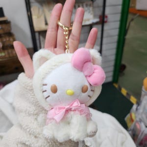 May include: White plush Hello Kitty keychain with a pink bow and a white fur coat. The keychain is attached to a gold chain.