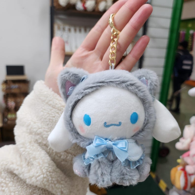 May include: A plush keychain featuring a white character with blue eyes and a pink nose wearing a gray hooded cat costume with a blue bow. The character has a small, white, fluffy tail.