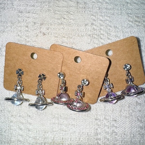 Può includere: Orecchini color argento con un design a pianeta, con globi trasparenti, rosa e viola. Ogni orecchino ha una piccola croce e una gemma scintillante. Gli orecchini sono esposti su cartoncini marroni per orecchini.