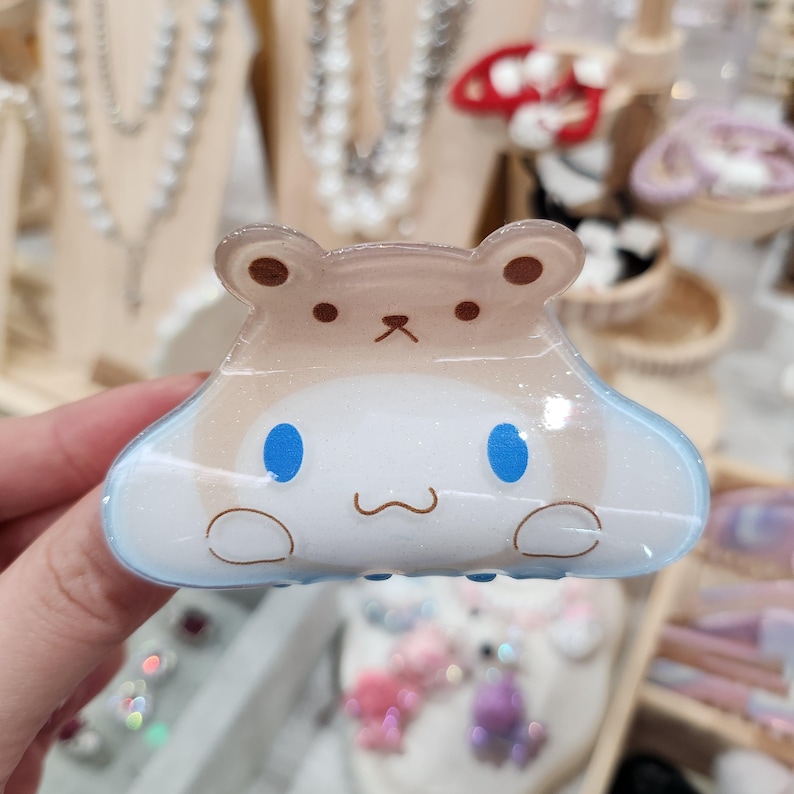 May include: A light blue and brown plastic hair clip shaped like a cartoon character with blue eyes and a white face. The character has a brown nose and a small mouth.