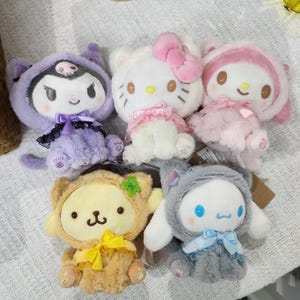 May include: Five plush toys in various colors and designs. The toys are all wearing outfits and have different facial expressions. The toys are sitting on a white surface.