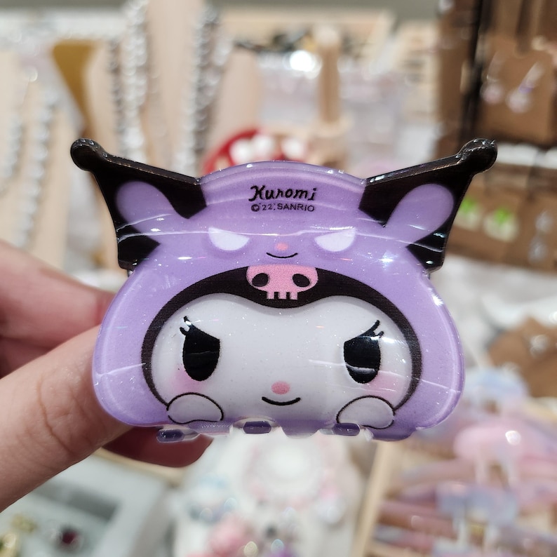 May include: Purple and black plastic hair clip featuring a cartoon character with a skull and crossbones design. The character is wearing a purple hooded outfit. The text "Kuromi &copy;'22 SANRIO" is printed on the clip.