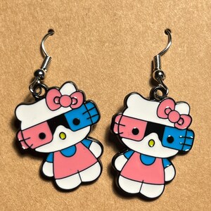 May include: A pair of silver Hello Kitty earrings with pink and blue 3D glasses. The earrings are made of metal and have a shiny finish.