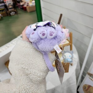 May include: A plush purple cat toy with a black bow and embroidered paw prints. The toy has a long tail and is held in a hand.