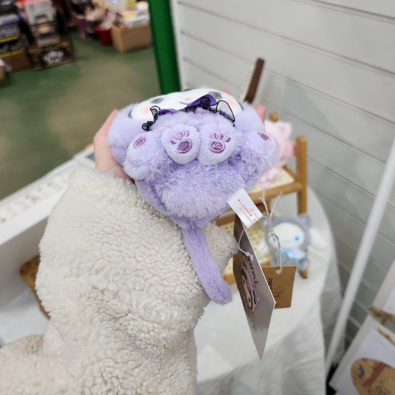 May include: A plush purple cat toy with a black bow and embroidered paw prints. The toy has a long tail and is held in a hand.