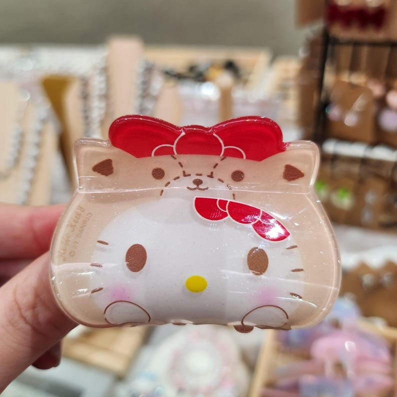 May include: A clear plastic hair clip with a Hello Kitty design. The clip features a red bow and a brown bear with a Hello Kitty face.