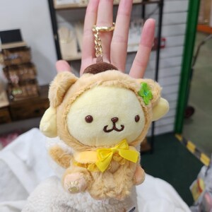 May include: A plush keychain featuring a yellow and brown dog wearing a brown and yellow costume with a yellow bow. The dog has a green flower on its head.