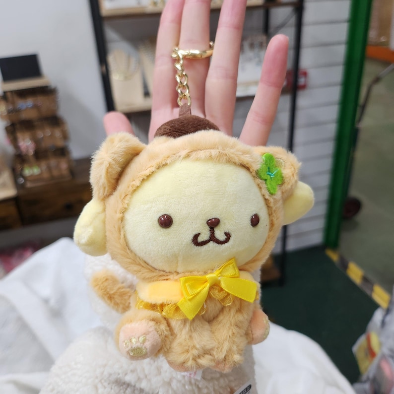 May include: A plush keychain featuring a yellow and brown dog wearing a brown and yellow costume with a yellow bow. The dog has a green flower on its head.