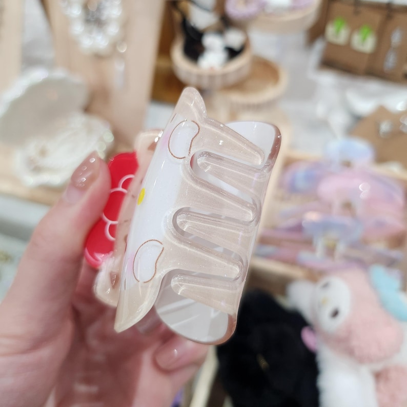 May include: A clear plastic hair claw with a red and white cartoon design. The claw has five teeth and is designed to hold hair in place.