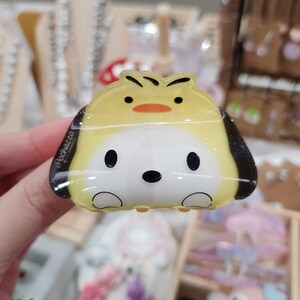 May include: A yellow and white plastic hair clip featuring a cartoon dog with a yellow chick on its head. The clip has the text "pochacco" on the side.