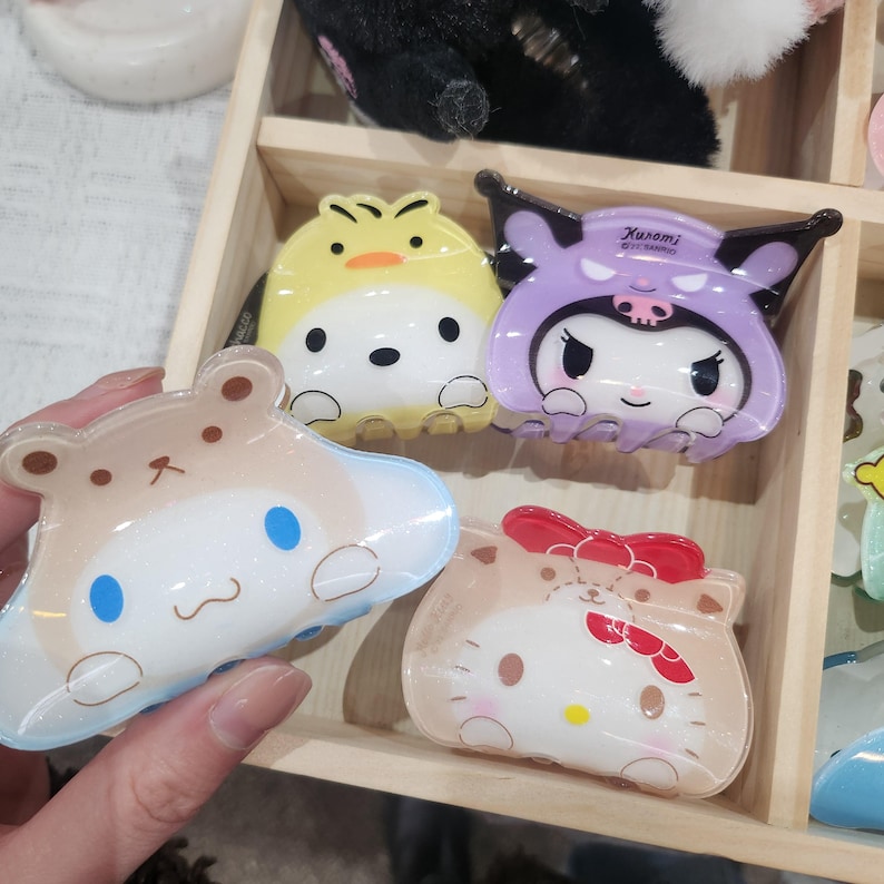 May include: Four colorful plastic hair clips featuring cartoon characters. The clips are shaped like a yellow chick, a purple bat with a skull and crossbones, a brown bear with blue eyes, and a pink cat with a red bow.