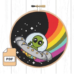 May include: Cross stitch pattern featuring a green alien in a spaceship flying through a rainbow in space. The pattern is in a round wooden embroidery hoop.