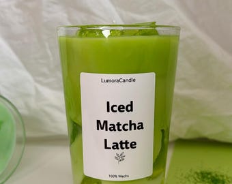 May include: A clear glass candle filled with green wax, resembling an iced matcha latte. The candle has a white label with the text "Iced Matcha Latte" and the brand name "LumoraCandle". The candle is decorated with green leaves and sits on a white and green surface.
