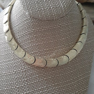 May include: A gold-toned necklace with a textured, wavy design. The necklace is made of linked, curved pieces that create a continuous, flowing pattern.