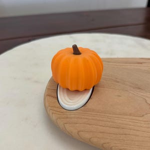 May include: An orange pumpkin-shaped bottle stopper with a brown stem sits on a wooden cutting board. The stopper is made of a smooth material and has a ribbed texture. The cutting board has a white ceramic insert.