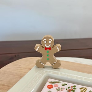May include: A tan gingerbread man figurine with a smiling face, red bow tie, and green buttons. The figurine has white icing details and is standing on a white tray with a festive holiday design.