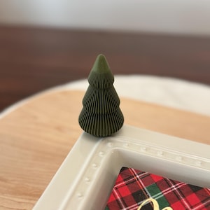 May include: A small, green, decorative Christmas tree with a layered design. The tree sits on a white tray with a red and green plaid card that has gold text. The tray is on a wooden surface.
