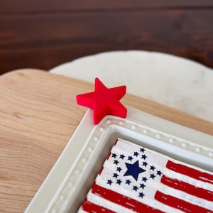 May include: White ceramic napkin holder with a red star on top. The napkin holder has a raised beaded edge and a design of an American flag inside.