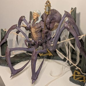 May include: A detailed fantasy figurine depicting a humanoid figure with long white hair, riding a large purple spider-like creature. The figure holds a lantern. The base is a stone-like structure with gold accents.