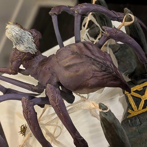 May include: A detailed sculpture of a fantastical creature, resembling a spider with a humanoid head. The figure is primarily purple with darker accents, and features intricate detailing. The head has white hair, and the sculpture is set against a neutral background.
