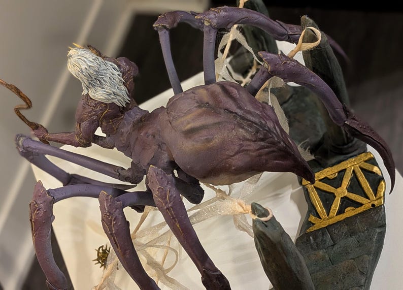 May include: A detailed sculpture of a fantastical creature, resembling a spider with a humanoid head. The figure is primarily purple with darker accents, and features intricate detailing. The head has white hair, and the sculpture is set against a neutral background.