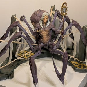 May include: A detailed figurine of a humanoid figure with long white hair, seated on a large, purple spider-like creature. The figure holds a lantern and is set against a backdrop of stone structures and faux spiderwebs. The color palette includes shades of purple, gray, and gold.