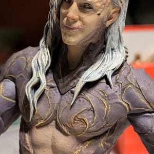 May include: A detailed figurine of a humanoid figure with pale skin, long white hair, and intricate armor. The figure has a stern expression and is adorned with gold accents. The chest and abdomen are exposed, revealing sculpted musculature.