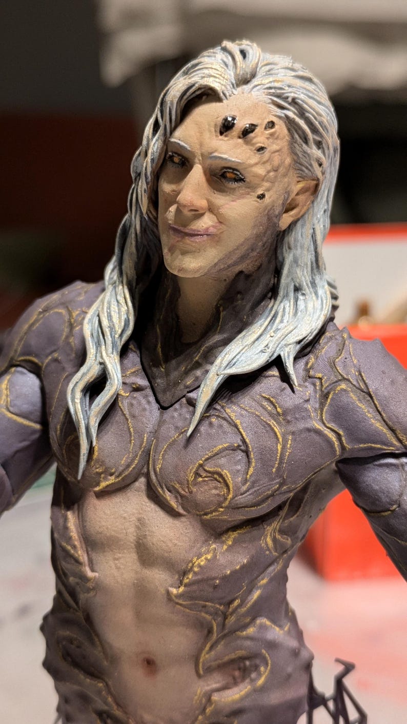 May include: A detailed figurine of a humanoid figure with pale skin, long white hair, and intricate armor. The figure has a stern expression and is adorned with gold accents. The chest and abdomen are exposed, revealing sculpted musculature.