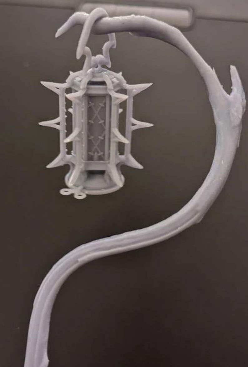 May include: A gray, ornate lantern with a spiky design, suspended from a curved, branch-like structure. The lantern features a detailed cage design and is likely a decorative item or a miniature model.