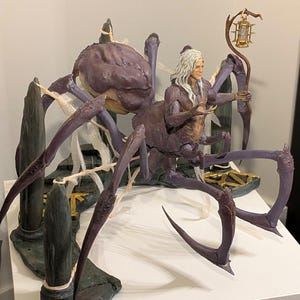 May include: A detailed figurine of a humanoid figure with long white hair, a purple spider body, and multiple legs. The figure holds a lantern and is set on a rocky base with a gold accent. The color palette includes shades of purple, gray, and gold.