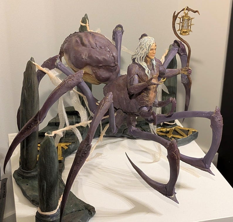 May include: A detailed figurine of a humanoid figure with long white hair, a purple spider body, and multiple legs. The figure holds a lantern and is set on a rocky base with a gold accent. The color palette includes shades of purple, gray, and gold.