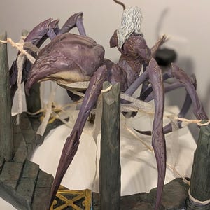 May include: A detailed fantasy sculpture of a large, purple spider with a rider. The spider has multiple legs and a textured body. The rider has long white hair and is wearing armor. The sculpture is set on a stone-like base with wooden posts.