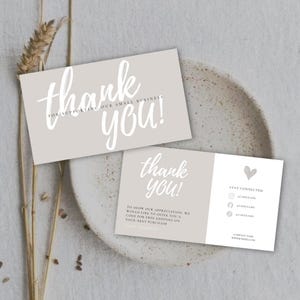 May include: Two light gray thank you cards with white script text. The cards say "thank you!" and "for supporting our small business." One card includes a heart and social media icons. The cards are on a neutral-toned plate.