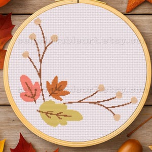 May include: A cross-stitch embroidery hoop with a fall-themed design featuring leaves and berries in shades of orange, pink, and green. The hoop is surrounded by autumn leaves, acorns, and a pumpkin on a wooden surface.