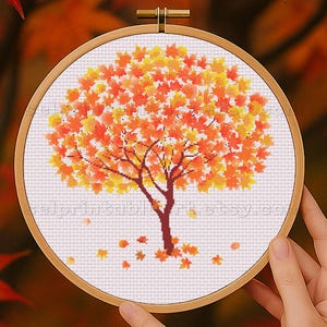 May include: A cross-stitch embroidery of a tree with orange and yellow leaves, set in a wooden hoop. The tree has a brown trunk and branches, with fallen leaves scattered below. The background is blurred with fall foliage.
