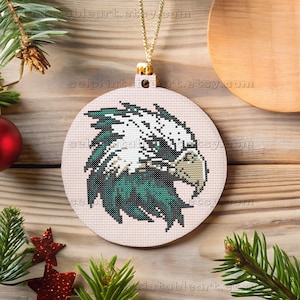 Può includere: Un ornamento natalizio rotondo con un disegno a punto croce della testa di un'aquila in verde, bianco e beige. L'ornamento ha un cappuccio dorato e un filo dorato per appenderlo. Lo sfondo è una superficie di legno con decorazioni festive.