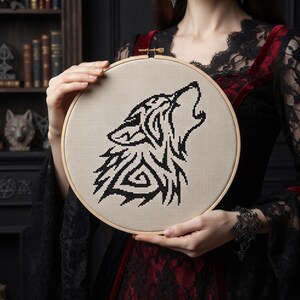 May include: A circular wooden embroidery hoop containing a piece of beige fabric with a black cross-stitch design of a howling wolf. The wolf design features geometric and tribal-inspired patterns throughout its silhouette, centered on the fabric.